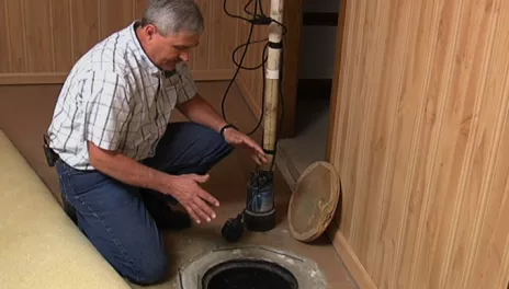 A man kneels beside a sump pump hole in the floor of a basement.