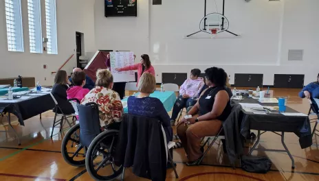 A group of people are gathered around a table in a gymnasium, watching a presenter point to a flipchart