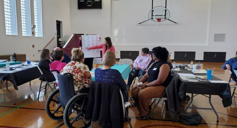 A group of people are gathered around a table in a gymnasium, watching a presenter point to a flipchart