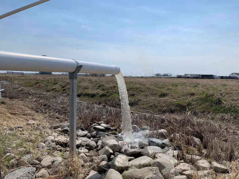 Water spills out of a white pipe onto large rocks at the edge of a ditch