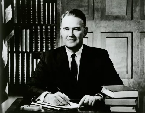 Quentin Burdick sits at a desk with American flag behind him to the right and bookshelves behind