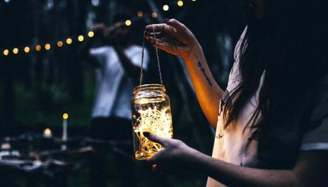 Girl holding a lit jar with a man string lights in a garden at night in the background
