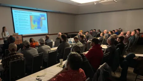 A speaker gestures toward a screen while addressing people in a dark room.