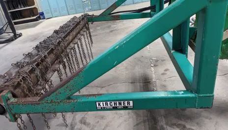 Green farm implement with metal frame holding a rotating drum with attached chains.