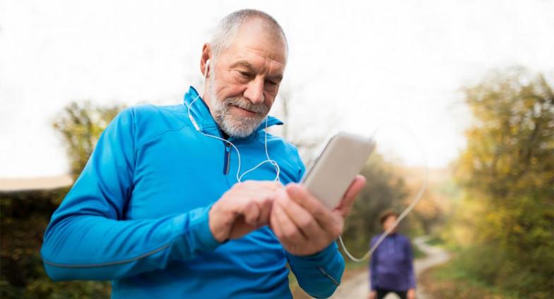 An older man standing on a running path looking at his mobile device. He's wearing athletic clothing and earbuds.