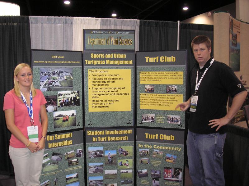two students pose in front of education poster for turf